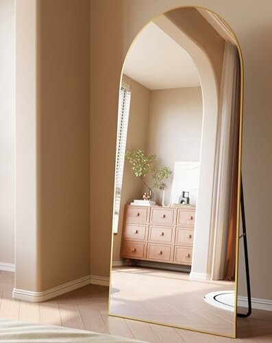 Large arched mirror reflecting a dresser with decor in a modern bedroom.